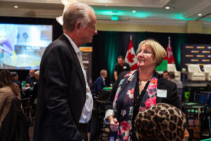 Brampton_Food and Bev Summit_Z62_9866 - Invest Brampton | Brampton Economic Development Two professionals converse at a conference, a man in a suit facing a woman with a colorful scarf and name badge. Behind them are flags and a presentation area.