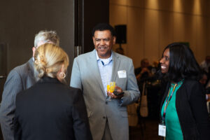 Brampton_Food and Bev Summit_Z62_9883 - Invest Brampton | Brampton Economic Development Professional networking moment: man in gray suit with name badge holds an orange drink while chatting with two women at a social event.