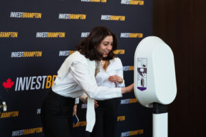 Brampton_Food and Bev Summit_Z62_9903 - Invest Brampton | Brampton Economic Development Woman in a white blouse operates a tall white kiosk with instructional diagrams, at an Invest Brampton backdrop.