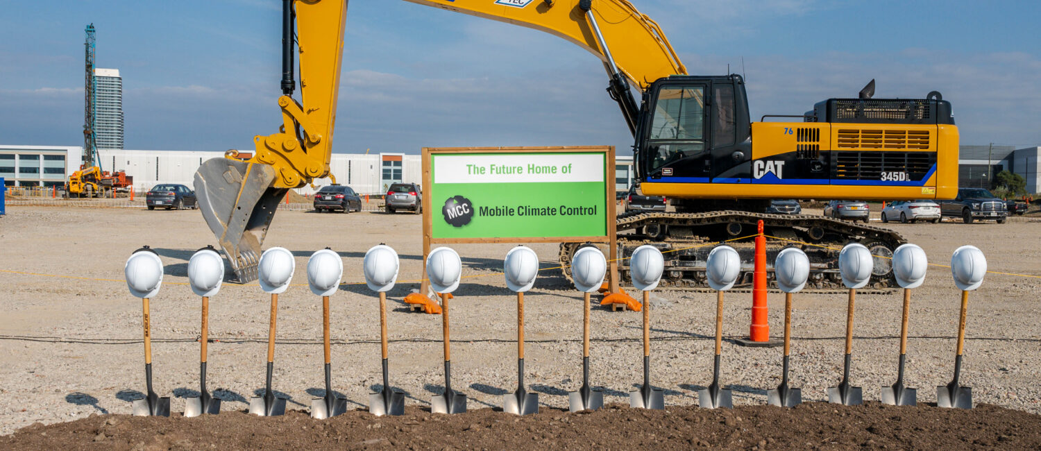 Yellow hydraulic excavator at a construction site, with a sign saying 'The Future Home of MCC Mobile Climate Control' and a row of safety helmets in the foreground.