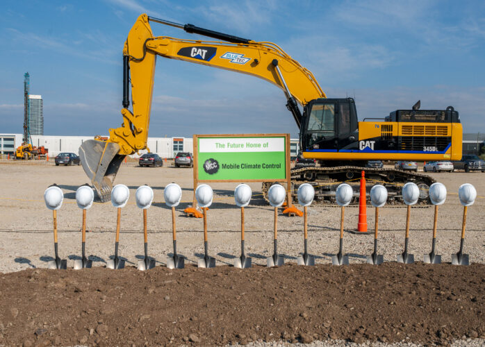 Yellow hydraulic excavator at a construction site, with a sign saying 'The Future Home of MCC Mobile Climate Control' and a row of safety helmets in the foreground.