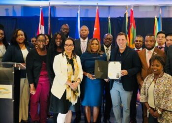 Group of diverse professionals posing on stage, one man holding a certificate, with flags in the background.