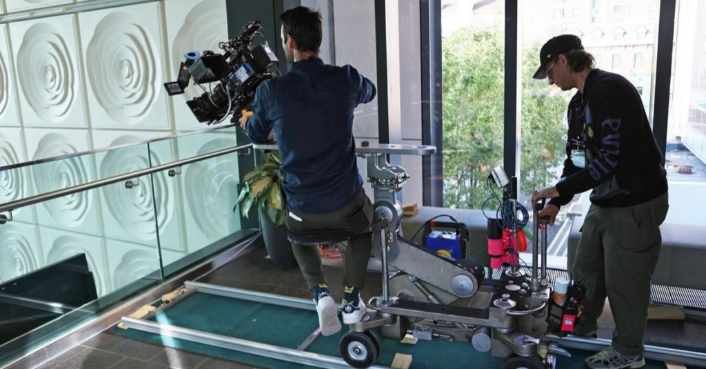 Cameraman operating a large cinema camera on a wheeled dolly track, with a crew member adjusting equipment near a glass railing inside a modern building.