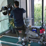 Cameraman operating a large cinema camera on a wheeled dolly track, with a crew member adjusting equipment near a glass railing inside a modern building.