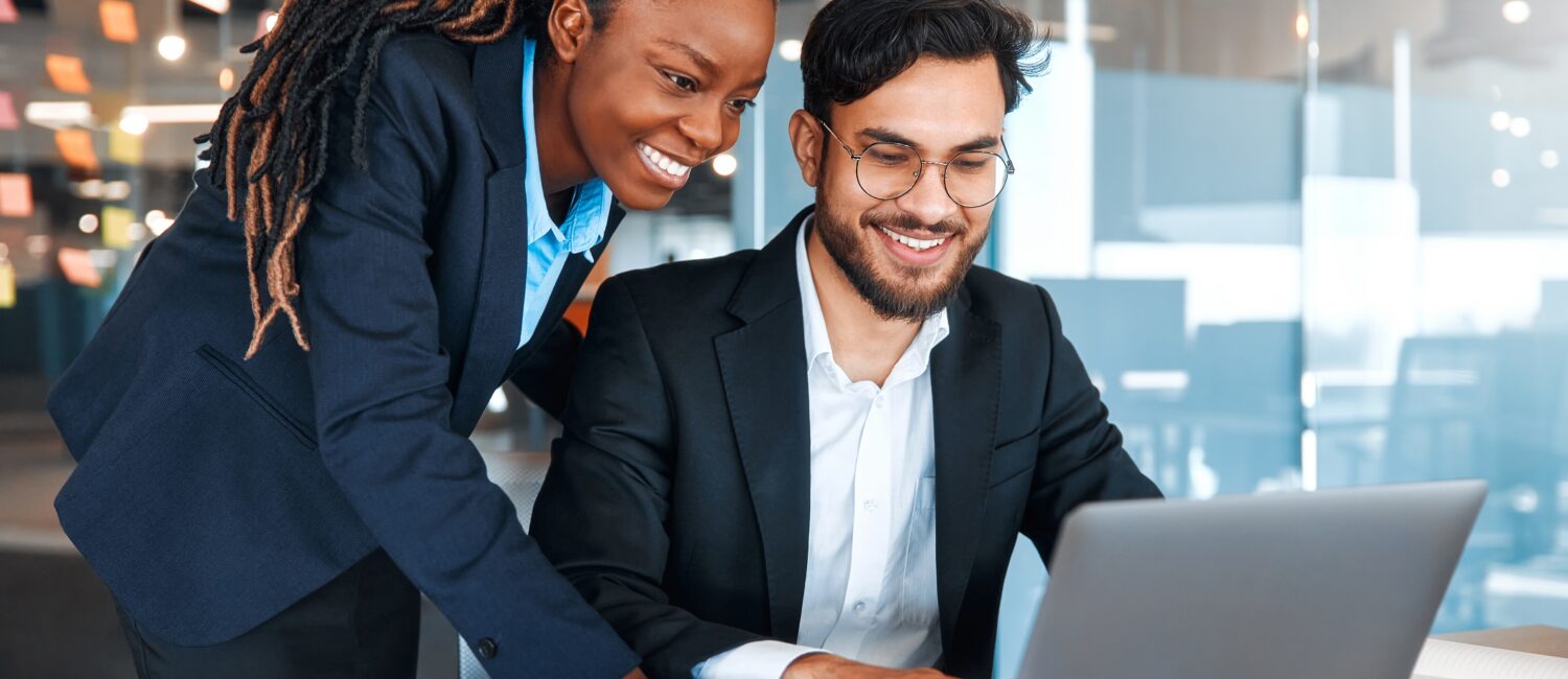 Two professionals in business attire collaborate over a laptop in a glass-walled office setting.