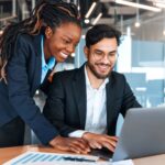 Two professionals in business attire collaborate over a laptop in a glass-walled office setting.
